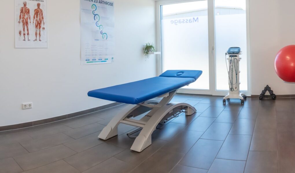 Blaue elektrische Therapieliege in einem hellen Behandlungsraum mit Anatomie-Postern und Physiotherapie-Geräten.
