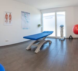 Blaue elektrische Therapieliege in einem hellen Behandlungsraum mit Anatomie-Postern und Physiotherapie-Geräten.