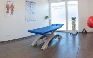 Blaue elektrische Therapieliege in einem hellen Behandlungsraum mit Anatomie-Postern und Physiotherapie-Geräten.