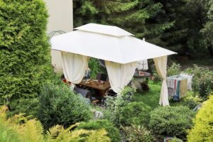 Gartenpavillon mit weissem Stoffdach und gemuetlicher Sitzgruppe inmitten dichter Buesche und Pflanzen