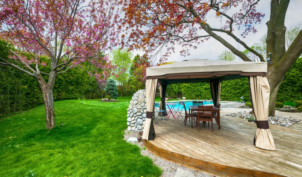 Moderner Gartenpavillon am Pool mit Holzdeck und eleganten Vorhaengen, umgeben von gepflegtem Rasen und Baeumen