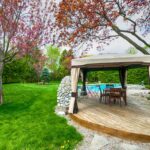 Moderner Gartenpavillon am Pool mit Holzdeck und eleganten Vorhaengen, umgeben von gepflegtem Rasen und Baeumen