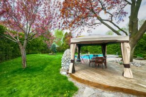 Moderner Gartenpavillon am Pool mit Holzdeck und eleganten Vorhaengen, umgeben von gepflegtem Rasen und Baeumen