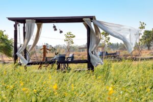 Offener Gartenpavillon aus Holz mit weissen Vorhaengen auf einer blumenreichen Wiese, natuerliche Gartengestaltung im Hintergrund